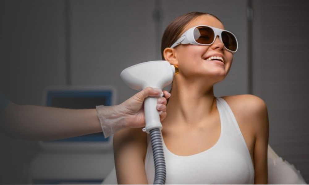 A woman smiling and wearing protective eyewear while receiving a laser hair reduction treatment on her face.