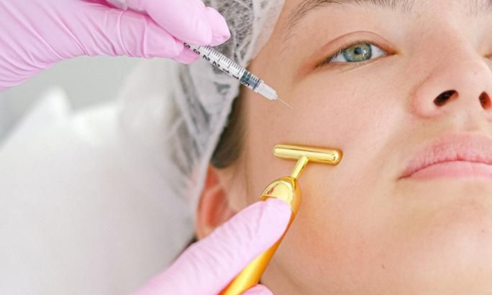 A woman is receiving two anti-aging cosmetic treatments simultaneously: a healthcare professional applies a gold T-shaped facial massager to her cheek while another person prepares an injection near her temple.