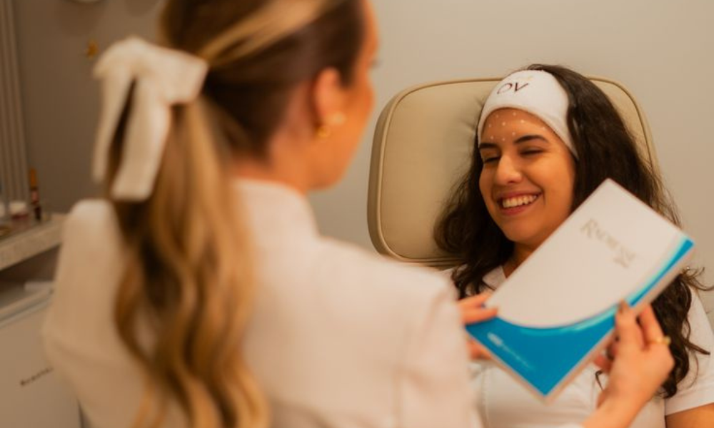 A smiling female client in a treatment chair wearing a white headband, looking towards a technician who is performing a treatment