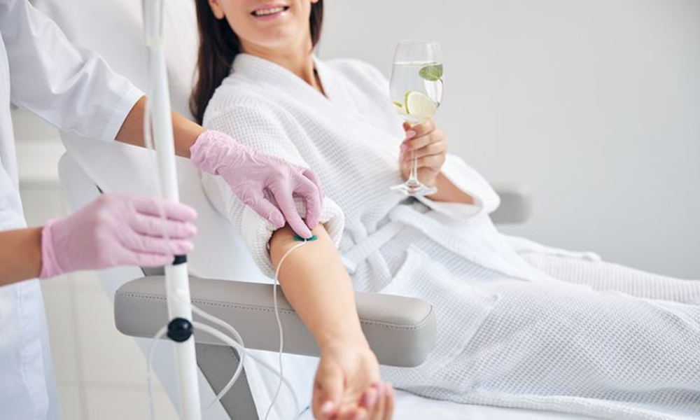 Person relaxing and smiling with an IV drip in their arm while holding a glass of water.